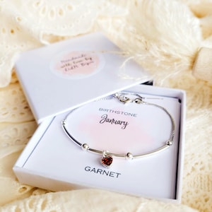 K&ouml;nnte beinhalten: Ein silbernes Armband mit einem herzf&ouml;rmigen Granat-Anh&auml;nger, pr&auml;sentiert in einer offenen wei&szlig;en Geschenkbox. Die Box enth&auml;lt eine Karte mit den Worten "Januar" und "Granat". Die Box hat einen Aufkleber mit der Aufschrift "Handmade with love by Edith Biju".