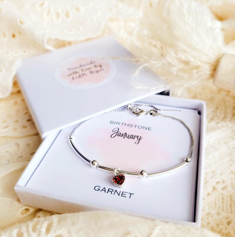 K&ouml;nnte beinhalten: Ein silbernes Armband mit einem herzf&ouml;rmigen Granat-Anh&auml;nger, pr&auml;sentiert in einer offenen wei&szlig;en Geschenkbox. Die Box enth&auml;lt eine Karte mit den Worten "Januar" und "Granat". Die Box hat einen Aufkleber mit der Aufschrift "Handmade with love by Edith Biju".