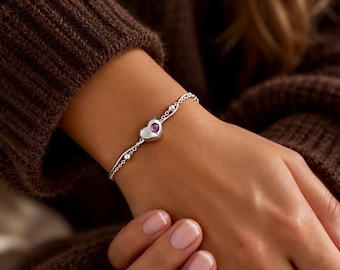 May include: A silver chain bracelet featuring a central heart-shaped charm with a small, round, dark pink gemstone set in the middle. The bracelet design includes delicate silver beads along the chain links, creating a refined and polished appearance.
