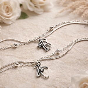 Peut inclure: Deux bracelets de cheville argentés avec des breloques initiales, portant les lettres "B" et "K". Chaque bracelet a un design de chaîne délicat avec de petites perles argentées. Le fond comprend des roses blanches et des accents floraux séchés.