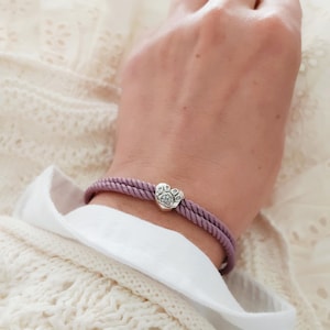 May include: A lilac rope bracelet featuring a silver paw print charm. The bracelet is worn on a wrist, set against a white sweater and shirt. The charm has a detailed paw print design.