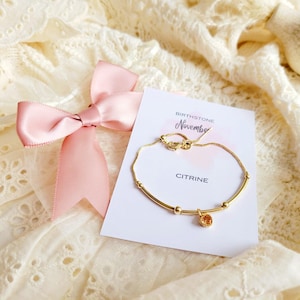 K&ouml;nnte beinhalten: Ein goldenes Armband mit einem Citrin-Edelstein-Anh&auml;nger, pr&auml;sentiert auf einer Karte mit der Aufschrift "Birthstone November Citrine". Eine rosa Schleife befindet sich ebenfalls im Bild, vor einem cremefarbenen Spitzenhintergrund.