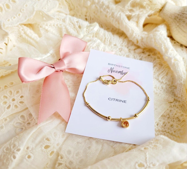 K&ouml;nnte beinhalten: Ein goldenes Armband mit einem Citrin-Edelstein-Anh&auml;nger, pr&auml;sentiert auf einer Karte mit der Aufschrift "Birthstone November Citrine". Eine rosa Schleife befindet sich ebenfalls im Bild, vor einem cremefarbenen Spitzenhintergrund.