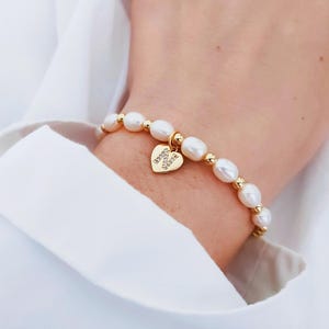 Peut inclure: Bracelet doré orné de perles d'eau douce et de perles dorées. Un charm en forme de cœur avec le texte "Happy Vibes" est attaché. Le bracelet est porté sur un poignet, sur fond blanc.