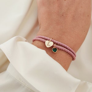 May include: A pink cord bracelet with two charms. One charm is a gold heart with the letter "L" and clear rhinestones. The other charm is a gold heart with a green gemstone.