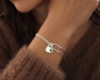 May include: Silver bracelet with a heart-shaped charm and a green gemstone. The bracelet has a delicate chain and a hinged design. The charm is a heart with a small, round green stone in the center.