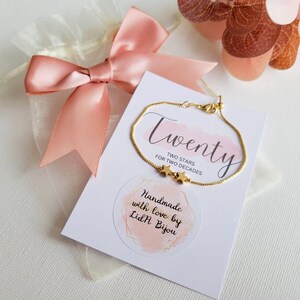 May include: A gold chain bracelet with two small star charms. The bracelet is displayed on a white card with the text "Twenty Two Stars for Two Decades" and "Handmade with love by Lidl Bijou".