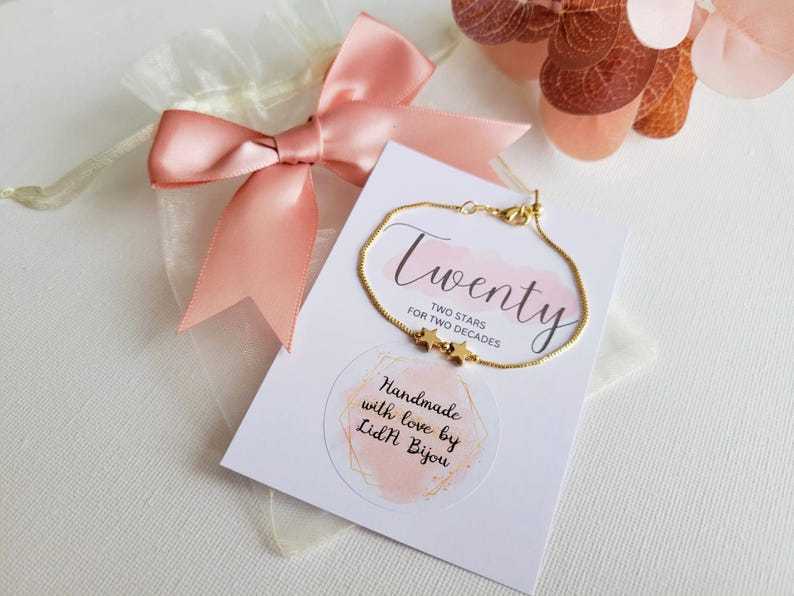 May include: A gold chain bracelet with two small star charms. The bracelet is displayed on a white card with the text "Twenty Two Stars for Two Decades" and "Handmade with love by Lidl Bijou".