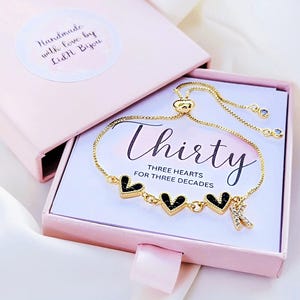 Könnte beinhalten: Ein verstellbares goldfarbenes Armband mit drei schwarzen Herzanhängern und einem Buchstaben-Anhänger K mit klaren Steinen. Das Armband liegt in einer hellrosa Geschenkbox mit einer Karte, auf der Thirty Three Hearts For Three Decades steht.