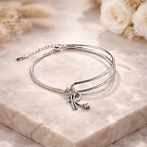 May include: A silver-toned bracelet with a double-band design and a chain closure. The bracelet features a charm with the letter "R" and a small, clear gemstone. The background includes white roses and dried plants.