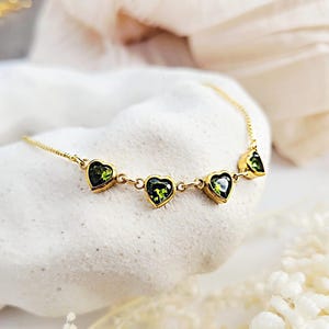 May include: A gold-colored necklace featuring five heart-shaped green gemstones. The necklace is displayed on a white, textured surface, with a soft, neutral-toned background. The gemstones are set in gold-colored bezels.