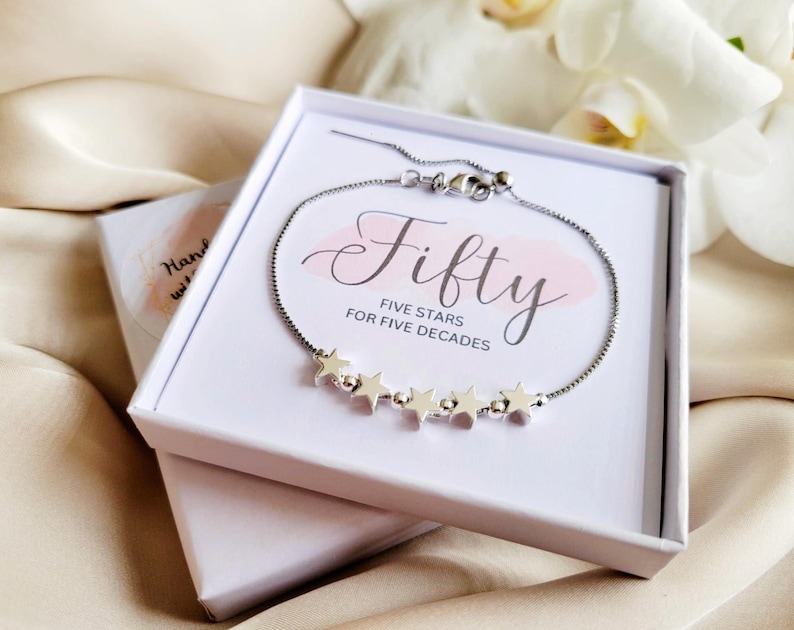 May include: A silver bracelet with five star charms in a white box. The box has the text "Fifty Five Stars for Five Decades" printed on it.