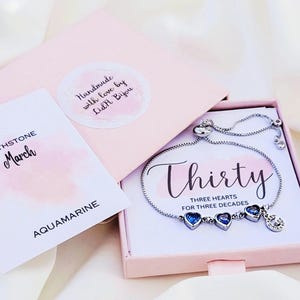 May include: A silver bracelet with three blue heart-shaped charms, presented in a pink box. The box also contains a card that says "BIRTHSTONE March AQUAMARINE". The bracelet is inscribed with the word "Thirty" and "THREE HEARTS FOR THREE DECADES".