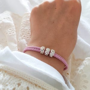 May include: A pink braided bracelet with three silver-colored, rhinestone-studded beads. The bracelet is worn on a wrist, against a backdrop of white fabric.