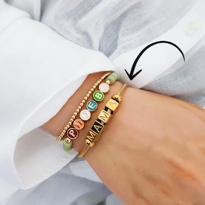 May include: Gold bracelets stacked on a wrist. One bracelet spells "MAMA" in black letters on gold cubes. Another bracelet has the name "PHOEBE" spelled out in colorful beads. A third bracelet is made of small gold beads.