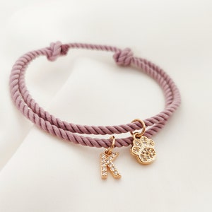 May include: A pink rope bracelet with a gold paw print charm and a gold letter K charm. The charms are encrusted with clear rhinestones.