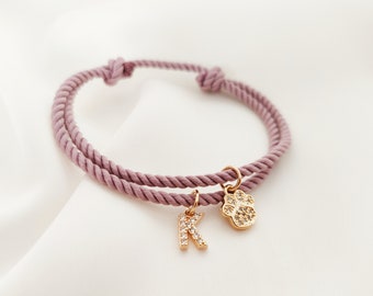 May include: A pink rope bracelet with a gold paw print charm and a gold letter K charm. The charms are encrusted with clear rhinestones.