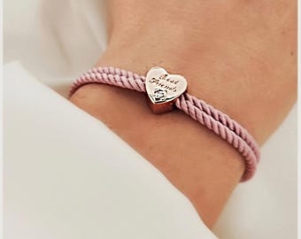 May include: A pink braided bracelet with a heart-shaped charm. The charm is rose gold with the words "Best Friends" and a small crystal. The bracelet is worn on a wrist.