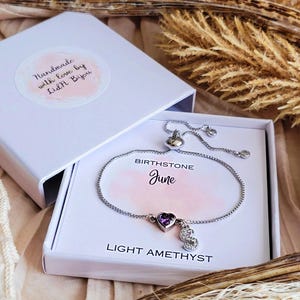 May include: A silver bracelet with a heart-shaped purple gemstone and a dangling letter "S" charm, presented in a white gift box. The box is labeled "Birthstone June" and "Light Amethyst". The box lid has a sticker that reads "Handmade with love by Lid&H Bijou".