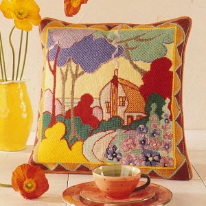 Può includere: Un cuscino in punto croce colorato che presenta una rappresentazione stilizzata di una casa con un tetto rosso, circondata da alberi e fiori. Il cuscino ha uno schema di colori giallo, verde, rosso e blu.