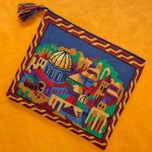 May include: An embroidered clutch bag with a vibrant cityscape design, including a golden-domed building. The design is framed by a dark blue border with a zig-zag pattern in red and gold. A multi-coloured tassel hangs from the top corner.