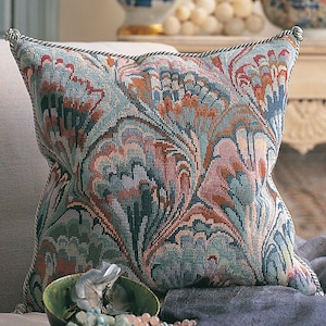 Puede incluir: Cojín decorativo cuadrado con un detallado estampado floral en tonos turquesa, coral y óxido. El cojín tiene un ribete de cuerda y está colocado sobre una superficie de color neutro. El diseño recuerda a un tapiz.