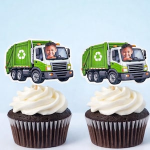 Pode incluir: Dois cupcakes com cobertura branca e bases de bolo de chocolate. Cada cupcake tem um topper decorativo de um caminhão de lixo verde e branco com uma criança motorista. O fundo é azul claro.