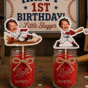 May include: Two clear glass mason jars filled with red crinkle paper, each topped with a wooden stick holding a cutout of a child in a baseball uniform. One cutout shows a child sliding into base, the other shows a child swinging a baseball bat.