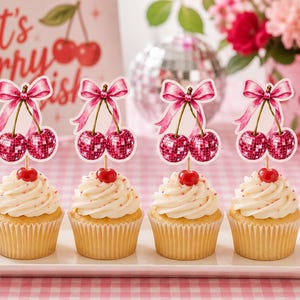 Op de afbeelding: Vier cupcakes op een rechthoekig wit bord, elk versierd met een roze discobal-kersen-topper met een strik. De toppers bestaan uit twee spiegelende, bolvormige kersen in een levendige roze kleur, verbonden door een roze lintje.