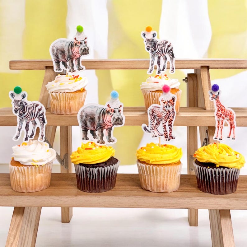 May include: Cupcakes with animal-themed toppers, including zebras, hippos, cheetahs, and giraffes, each wearing a party hat. The cupcakes are arranged on a wooden tiered stand, with yellow and white frosting.