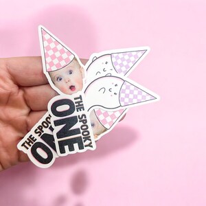 May include: A set of die-cut stickers featuring a baby with a surprised expression wearing a pink and white checkered party hat, alongside ghost illustrations with matching hats. The text reads THE SPOOKY ONE in bold black and white lettering.