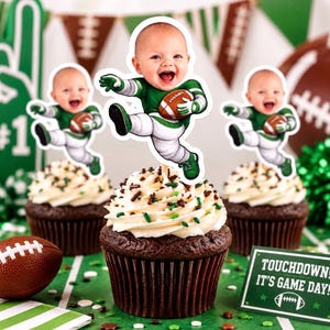 May include: Three chocolate cupcakes with white frosting and sprinkles, each topped with a custom paper cutout of a baby wearing a green and white football uniform and holding a football. A small sign reads TOUCHDOWN! IT'S GAME DAY!