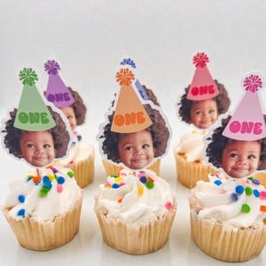 Puede incluir: Cupcakes con glaseado blanco y chispas de colores, cubiertos con recortes de la cara de un ni&ntilde;o que lleva sombreros de fiesta que dicen "ONE". Los sombreros son de varios colores, incluyendo verde, morado, naranja y rosa.