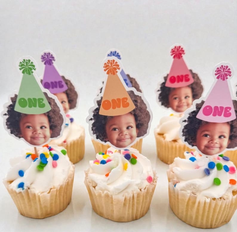 Puede incluir: Cupcakes con glaseado blanco y chispas de colores, cubiertos con recortes de la cara de un ni&ntilde;o que lleva sombreros de fiesta que dicen "ONE". Los sombreros son de varios colores, incluyendo verde, morado, naranja y rosa.