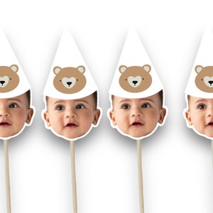 May include: Four white paper party hats with brown bear faces and a baby's face peeking out from the bottom of each hat. The hats are attached to wooden sticks.
