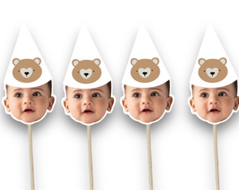 May include: Four baby face cutouts with white party hats featuring a brown bear face design. The cutouts are attached to wooden sticks, perfect for a baby shower or birthday party.