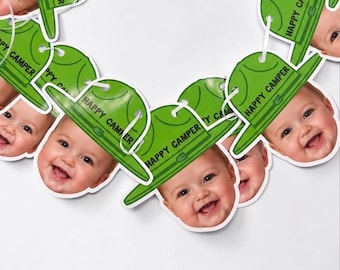 May include: A decorative garland featuring multiple cutouts of smiling infants wearing green hats. Each hat is printed with the text HAPPY CAMPER. The cutouts are connected by a thin white string, creating a repeating pattern of faces and hats.