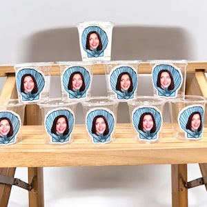 May include: Clear plastic shot glasses with a seashell design and a woman's face printed on them. The seashell is light blue, and the woman has dark hair. The shot glasses are arranged on a wooden bench.