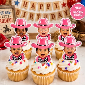 May include: Six cupcakes topped with white frosting and colorful sprinkles, each featuring a custom paper topper of a smiling baby wearing a bright pink cowboy hat and a black and white cow-print bandana. The toppers have a glossy finish.