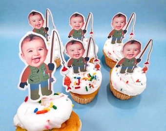 Könnte beinhalten: Cupcakes mit weißem Zuckerguss, bunten Streuseln und Baby-Angelruten-Toppern. Die Topper zeigen ein lächelndes Baby in einer Angelweste, das eine Angelrute mit einem Fisch hält. Die Cupcakes stehen auf einer hellblauen Oberfläche.