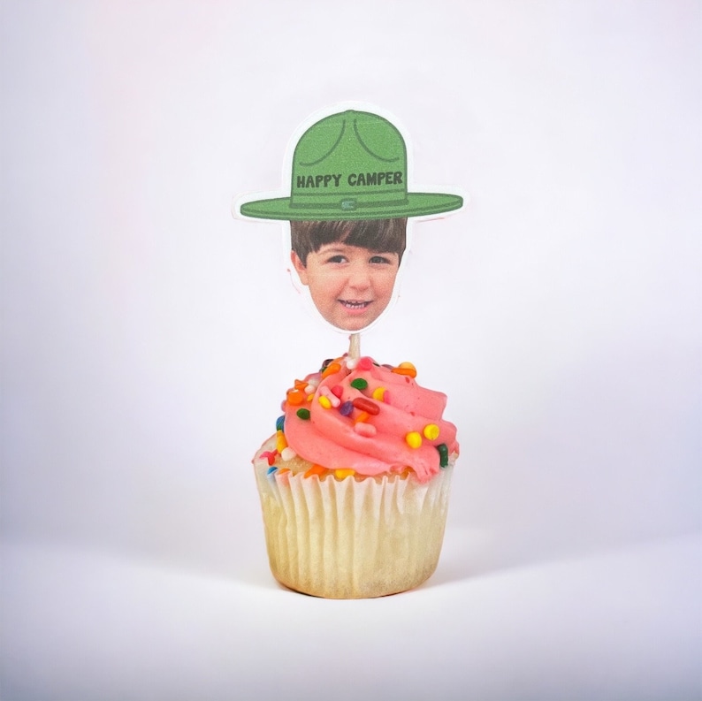 Op de afbeelding: Een cupcake met roze glazuur en kleurrijke sprinkles, bekroond met een foto van een kind met een groene hoed met de tekst "HAPPY CAMPER". De cupcake zit in een witte papieren vorm.