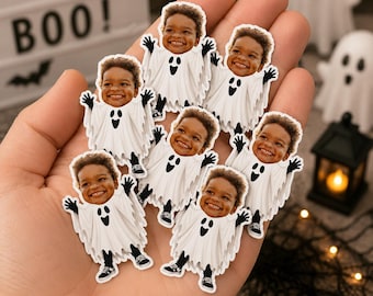 May include: A collection of small, die-cut stickers featuring a smiling child's face superimposed onto a white ghost costume with black details. Each sticker shows the child with arms raised, wearing black shoes, and a ghost sheet with a black face design.