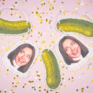May include: A festive garland featuring green pickle cutouts and smiling woman's face cutouts, strung together with white string. The background is a light pink with gold confetti scattered throughout.