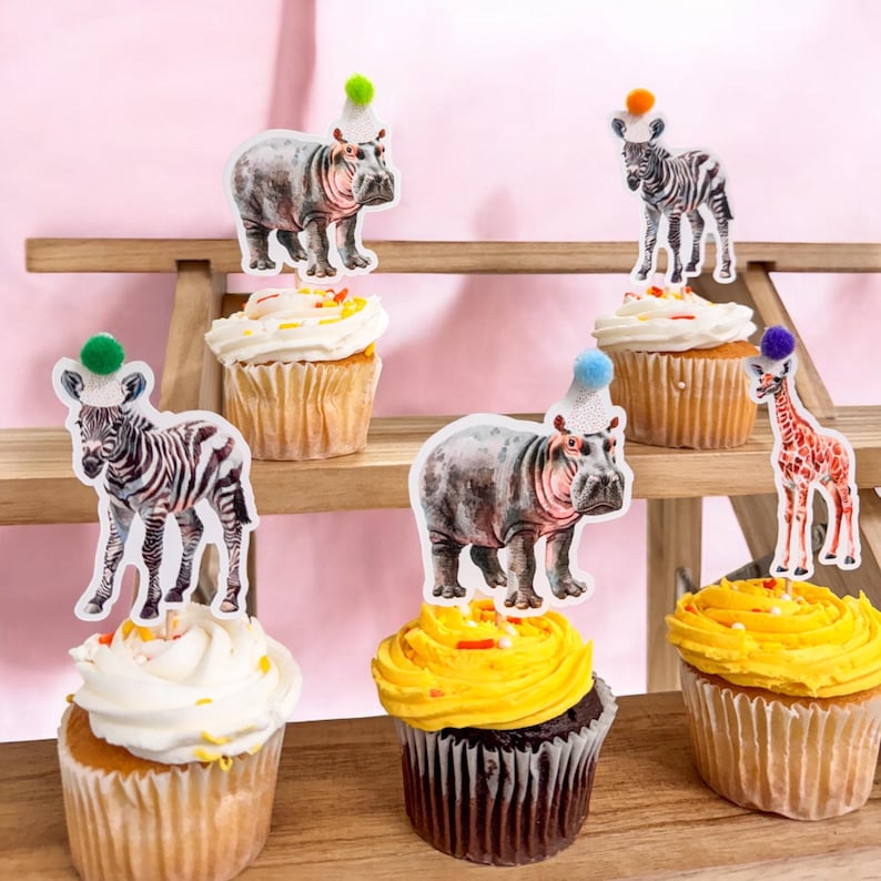 May include: Cupcakes with animal-themed toppers, including zebras, hippos, and a giraffe, each wearing a party hat with a colorful pom-pom. The cupcakes have white and yellow frosting, and are arranged on a wooden shelf against a pink background.