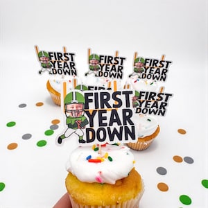 May include: Cupcakes with white frosting and colorful sprinkles, topped with football-themed toppers. The toppers feature a cartoon football player and the text "FIRST YEAR DOWN" in black and white.