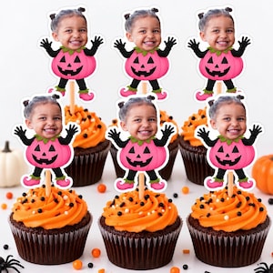 May include: Six cupcake toppers featuring a cutout of a smiling child's face attached to a pink pumpkin costume with black sleeves and leggings, mounted on wooden sticks and placed into orange frosted cupcakes with black and white sprinkles.