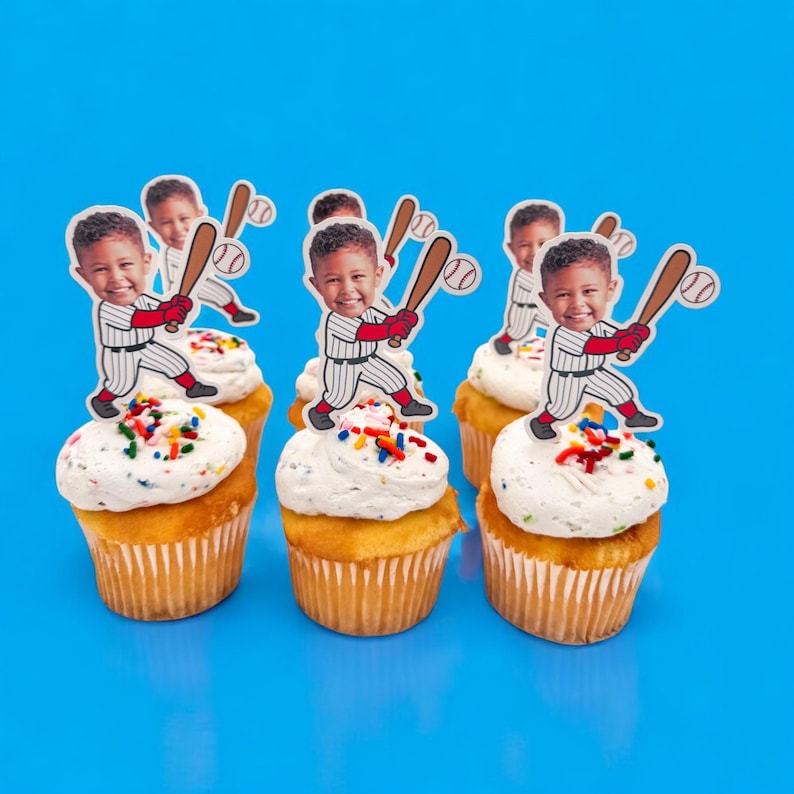 May include: Six cupcakes with white frosting and colorful sprinkles. Each cupcake is topped with a baseball-themed cutout featuring a child holding a bat. The background is a solid bright blue.