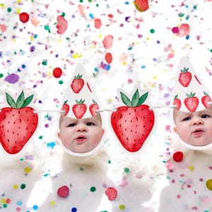 May include: A white banner with red and green strawberries and a baby's face in each section. The banner is decorated with confetti.