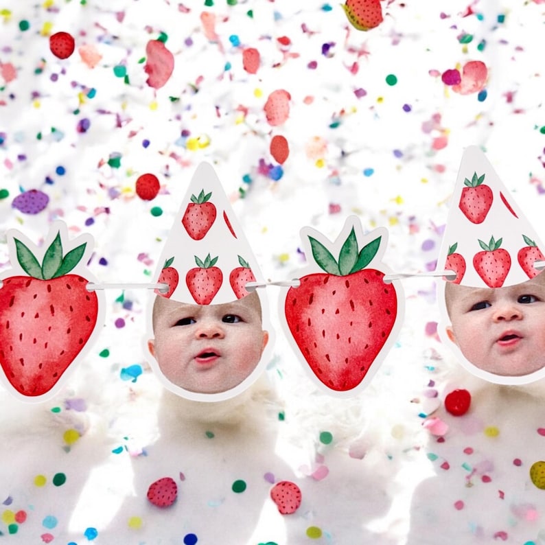 May include: A white banner with red and green strawberries and a baby's face in each section. The banner is decorated with confetti.