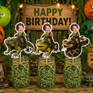 May include: Three decorative cake toppers featuring a child's face on a lizard, a frog, and a snake, each mounted on a stick inside a glass mason jar filled with green filler. The background includes balloons, a wooden sign reading HAPPY BIRTHDAY.